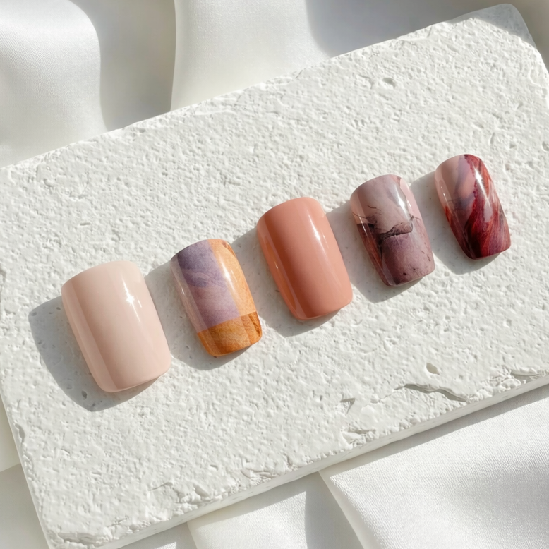 Five press-on nails with warm terracotta, mauve, and marble swirl designs arranged on a white stone tile.