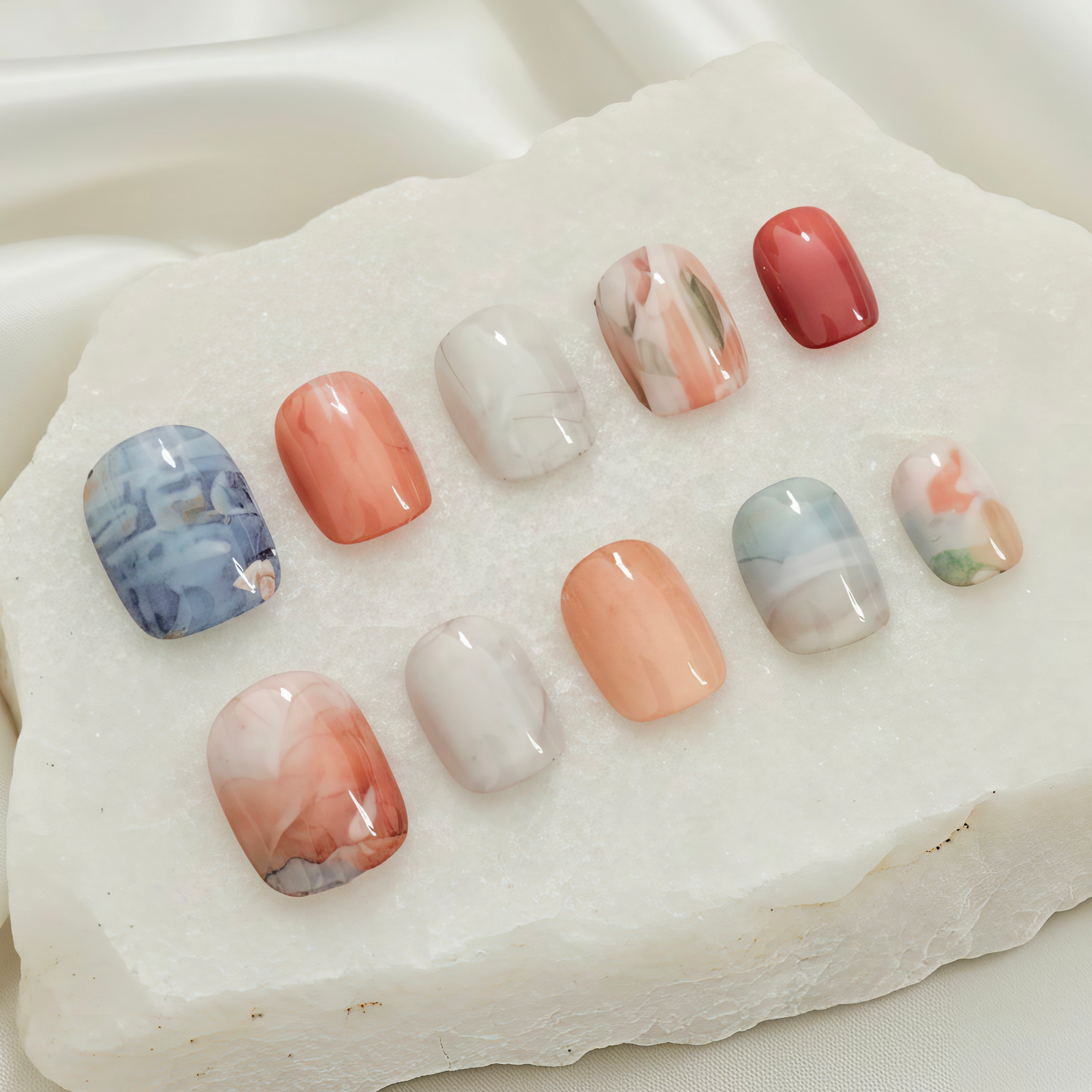 A collection of ten press-on nails with varied marble, swirl, and solid coral-orange designs in a stone display.