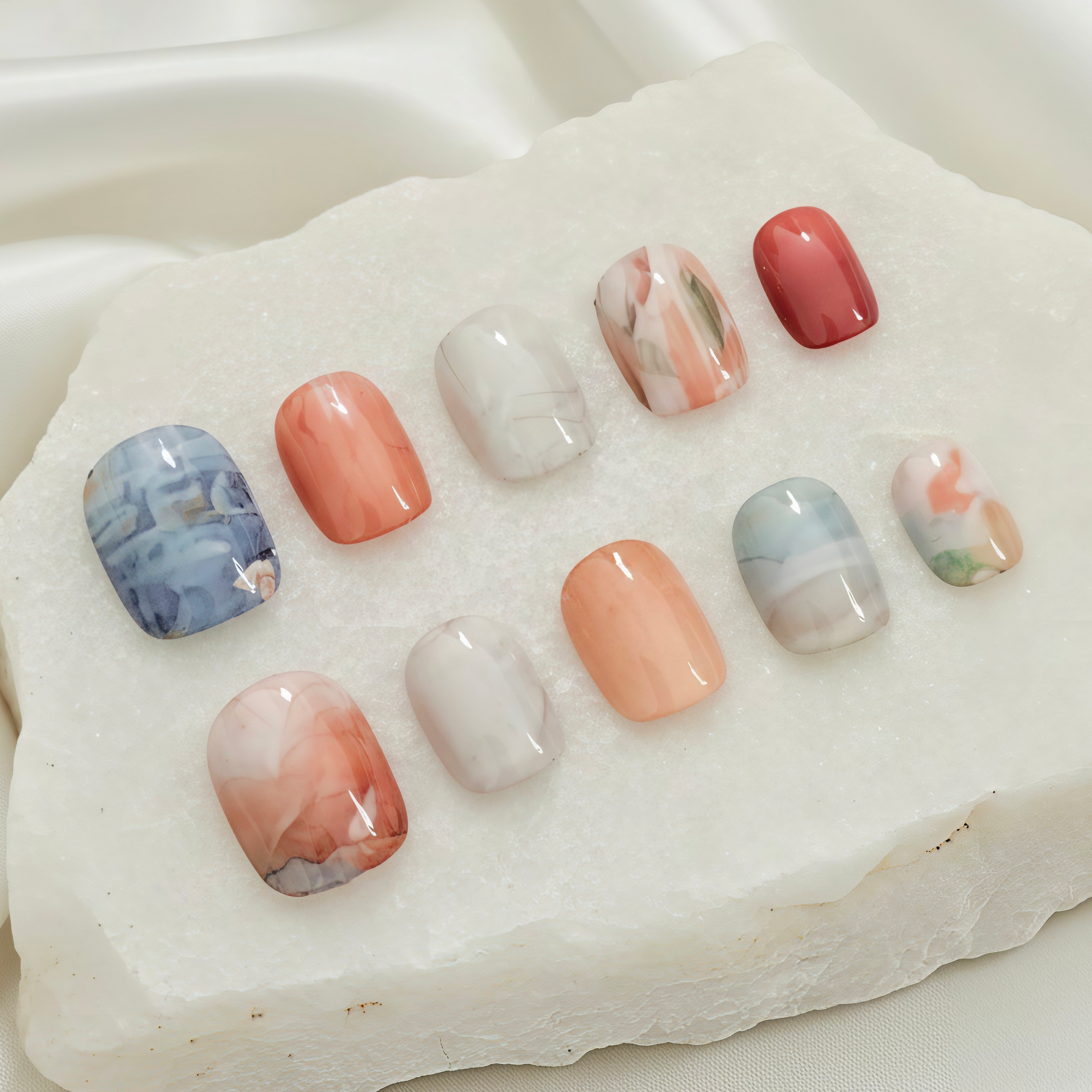 A collection of ten press-on nails with varied marble, swirl, and solid coral-orange designs in a stone display.