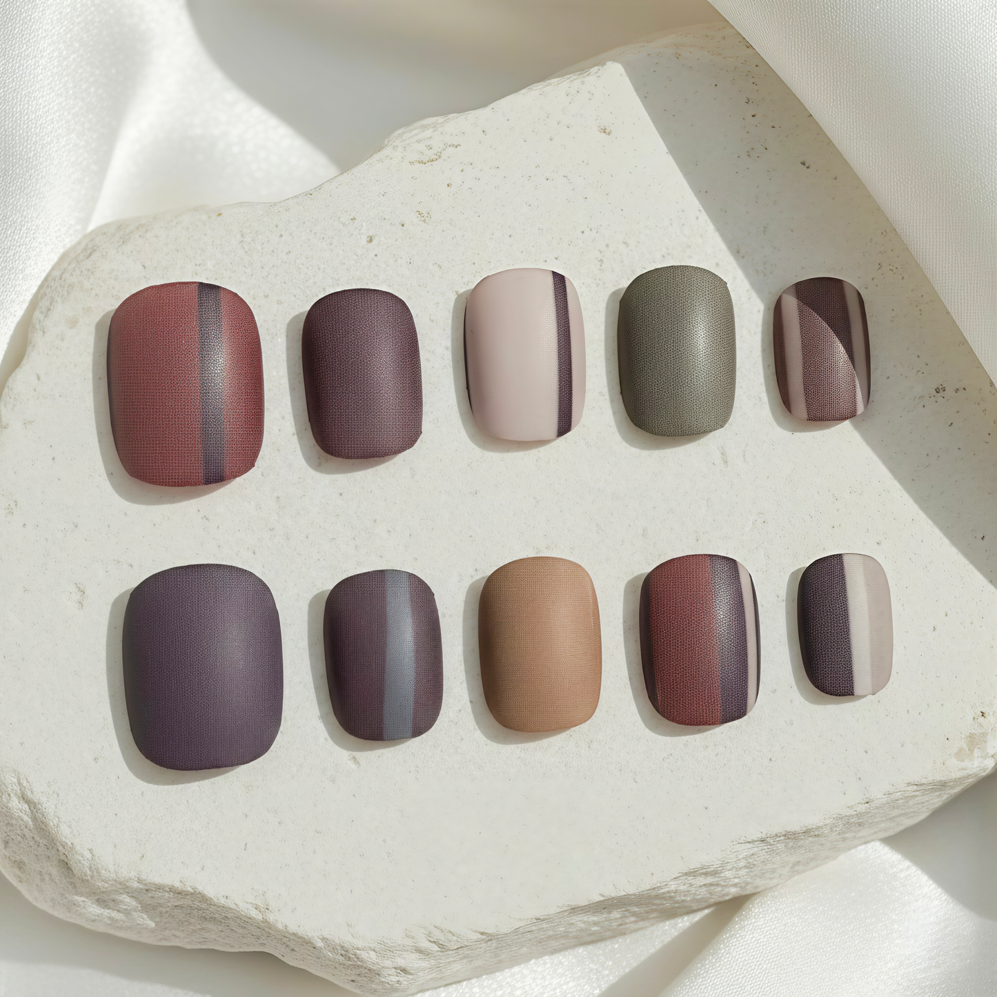 A set of ten textured press-on nails in deep plum, slate, and tan with minimalist vertical stripe accents.